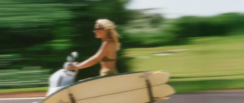 Woman riding vintage scooter with surfboard along tropical coast